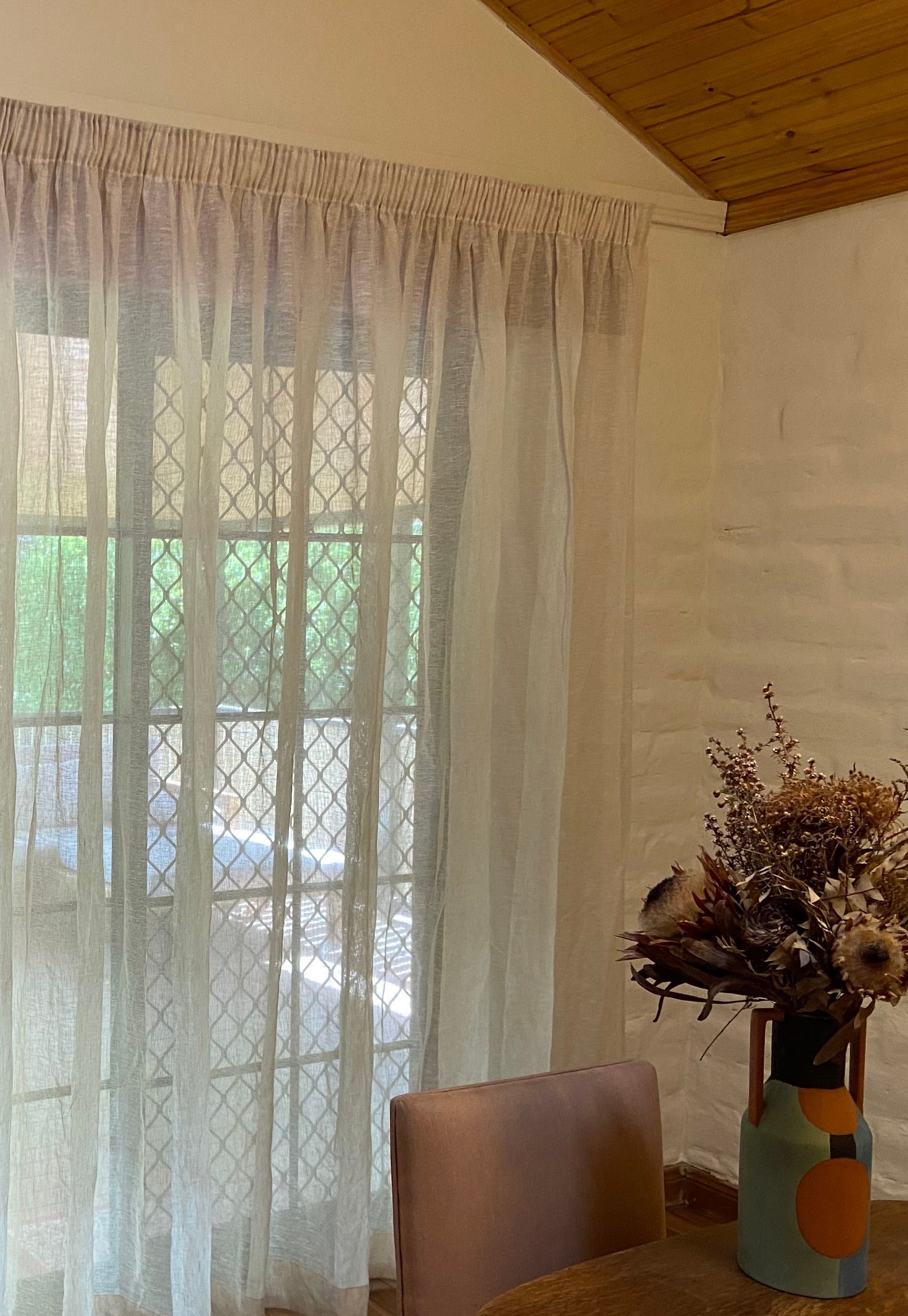 Sheer white curtains hang in front of a window, with a vase of dried flowers on a table in the foreground.