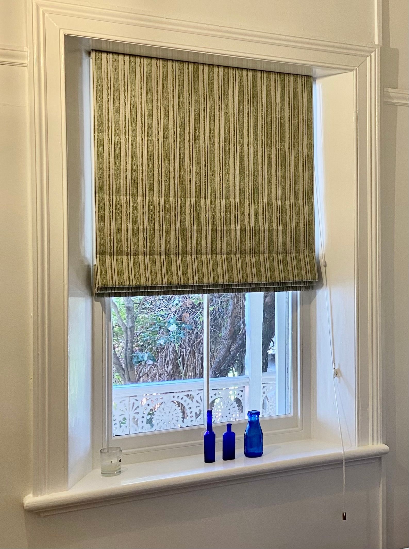 A window with a green and white patterned Roman shade, featuring three blue glass bottles on the sill.