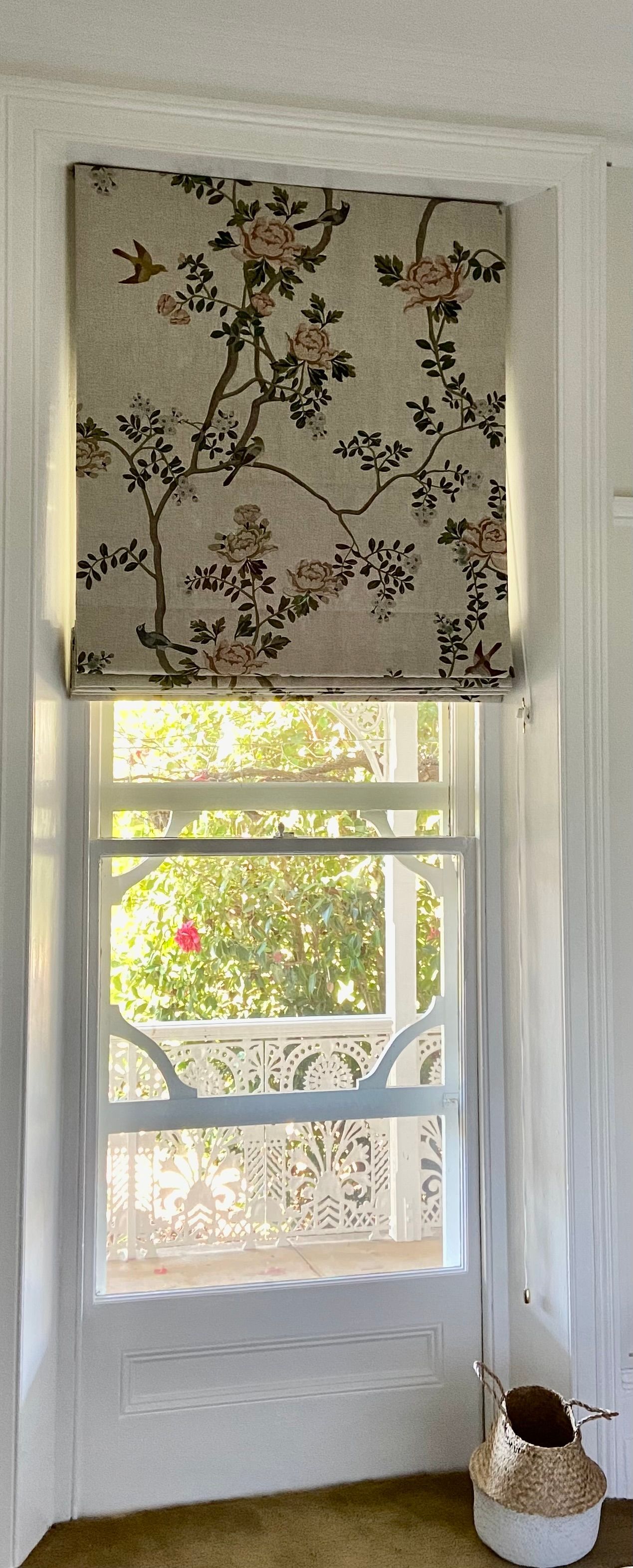 A window with a floral Roman shade above a white-framed view of a porch with decorative lattice railings.