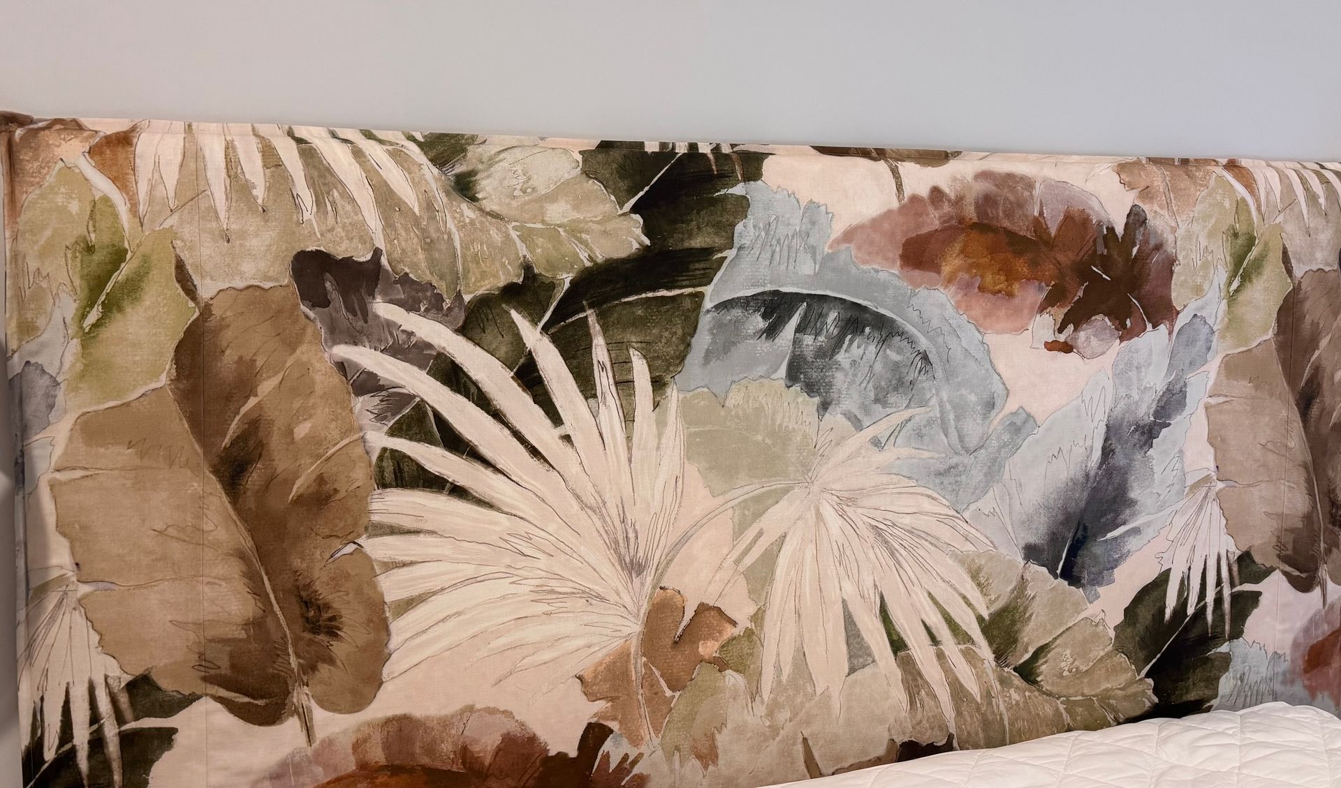 A headboard upholstered in fabric featuring a botanical print of large palm leaves in shades of tan, brown, and sage green.