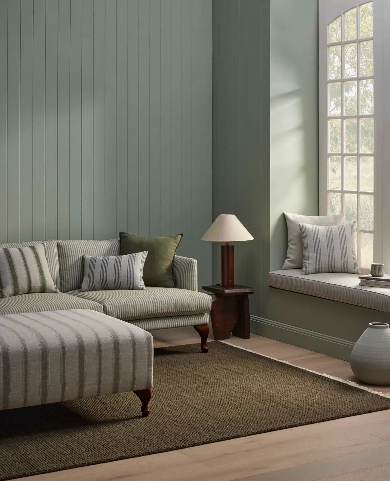 A sage green room with a striped sofa, matching ottoman, a wooden lamp on a side table, and a window seat with pillows.