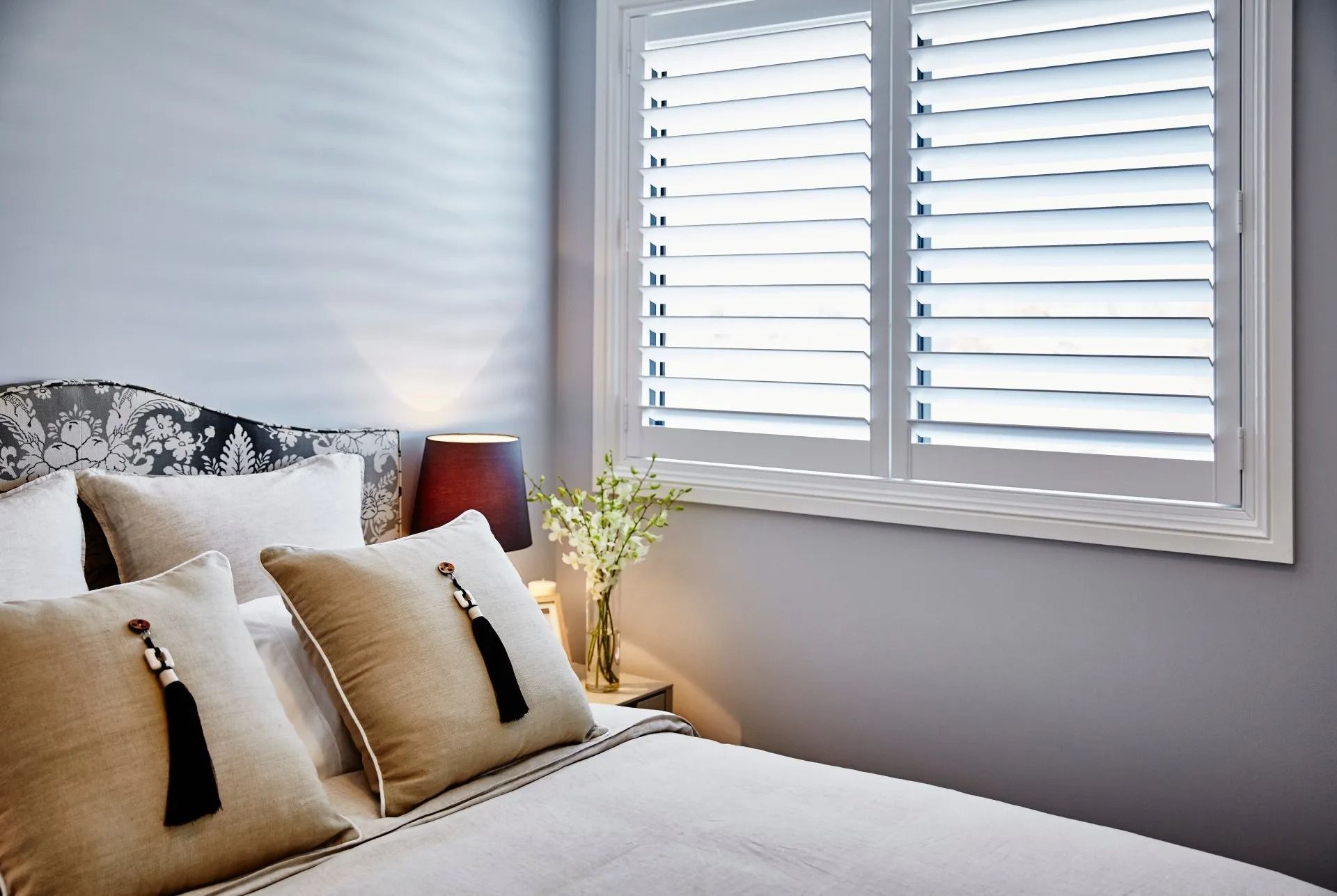 Bedroom With White Shutters, Bed With Pillows, and a Nightstand With a Lamp and Flowers — Red Door Decor In Dubbo, NSW