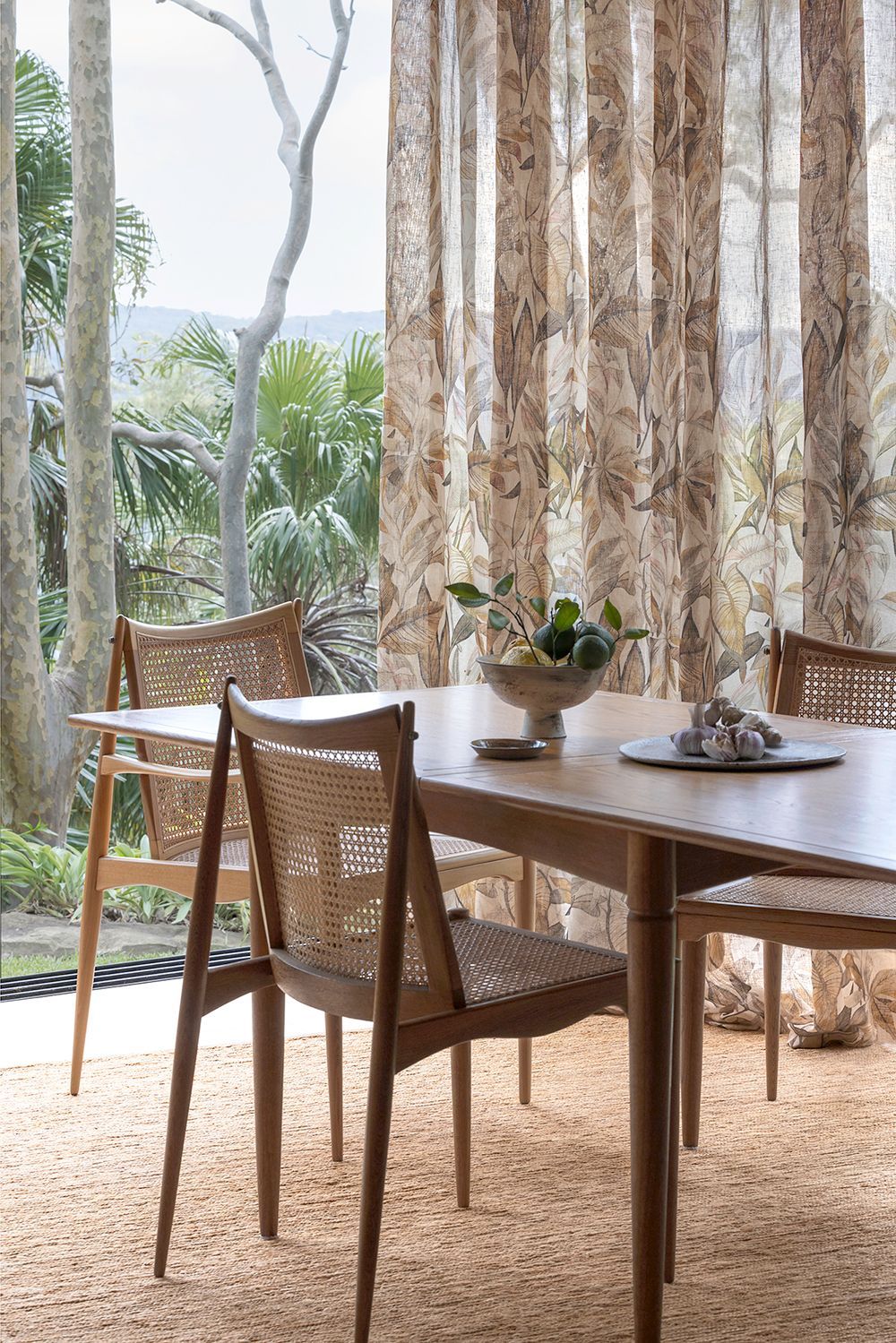 Dining room with a wooden table, woven chairs, and floral curtains, overlooking trees.