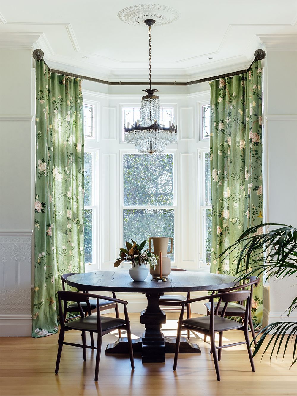 Dining room with a round wooden table, chairs, and green floral curtains around a large window.