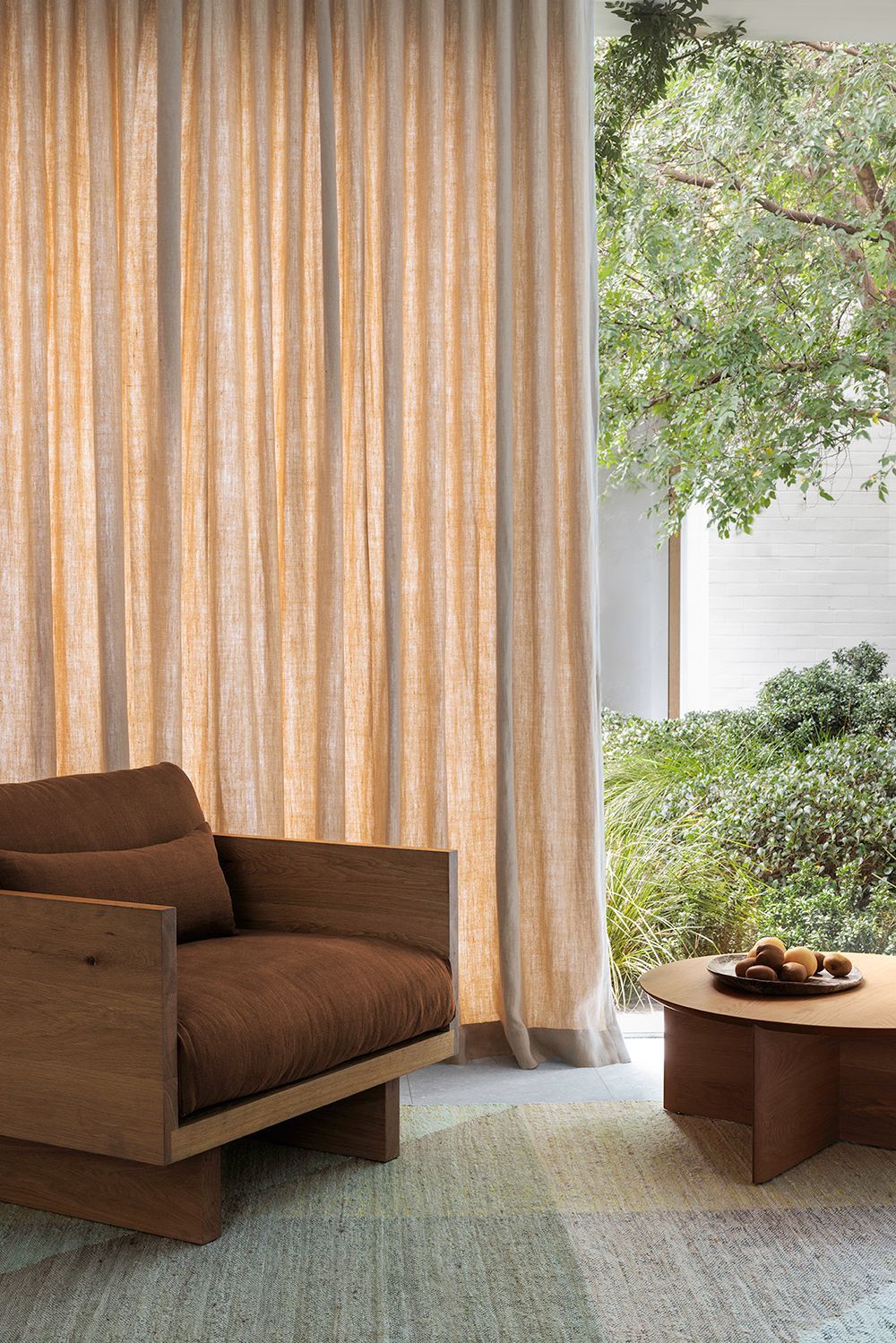 Cozy living room with brown chair, wooden coffee table, and beige curtains framing a view of greenery.