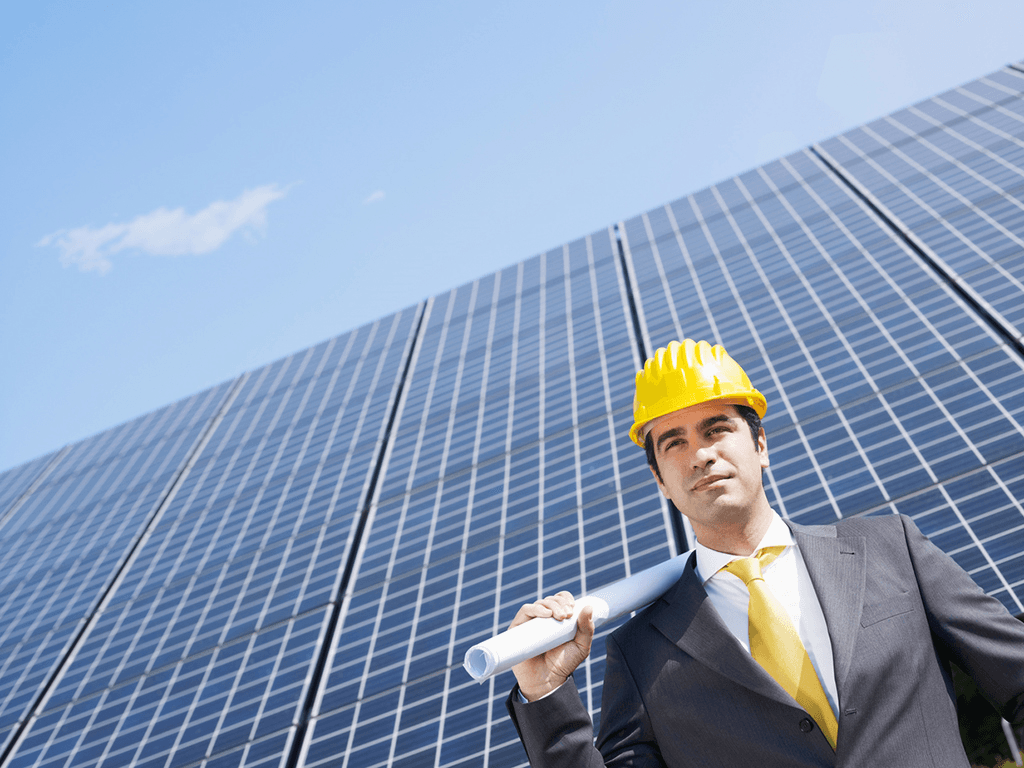Businessperson in a yellow hard hat holding blueprints in front of solar panels