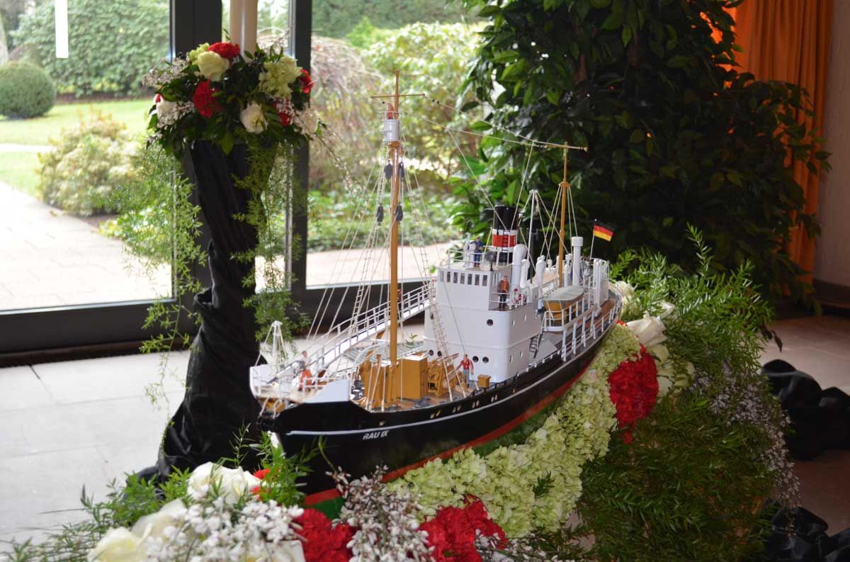 Auf einem Tisch steht ein Modellboot, umgeben von Blumen.