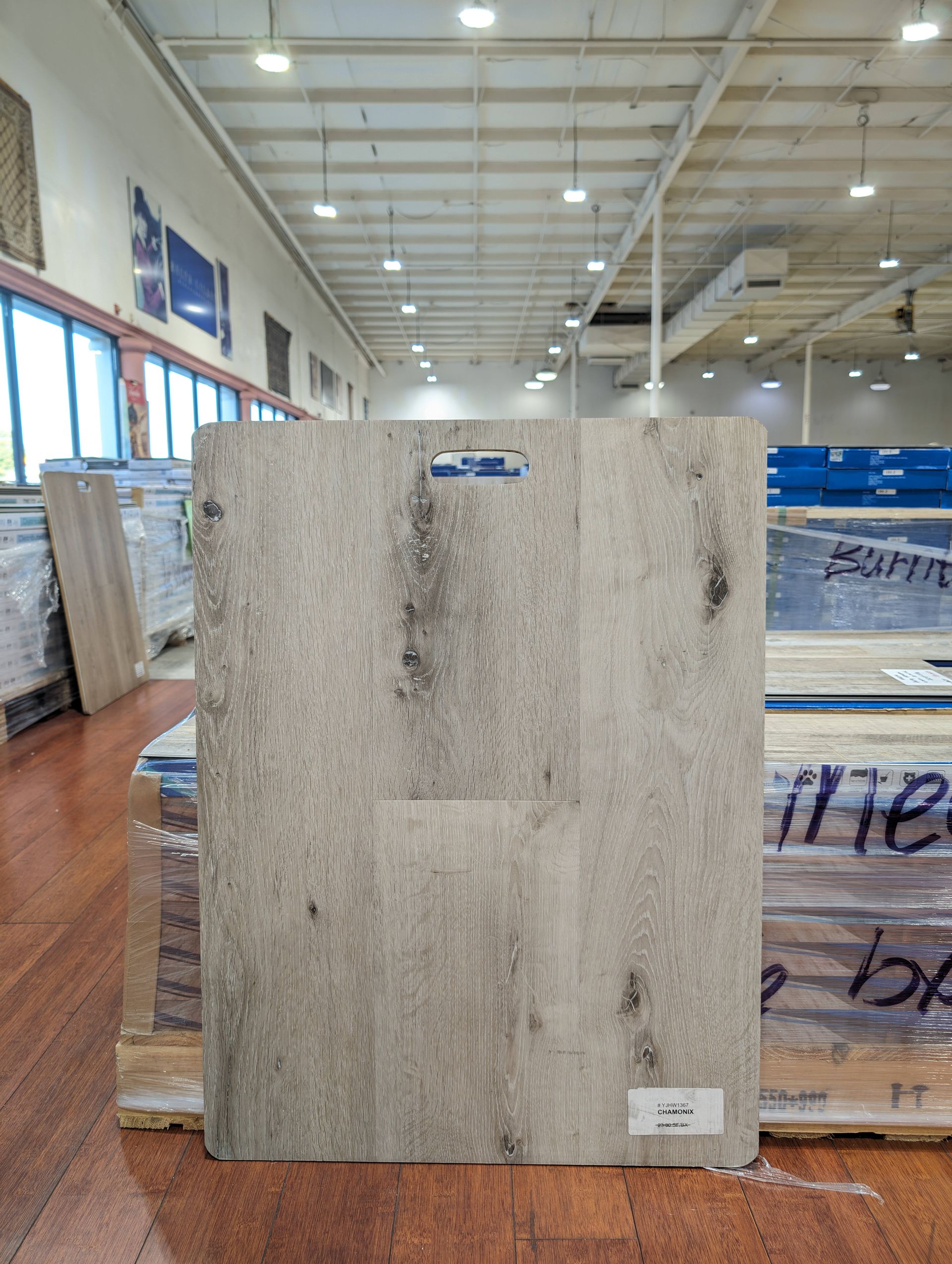 Wooden floorboard sample, light gray with dark streaks, indoors on a wood pallet in a warehouse.