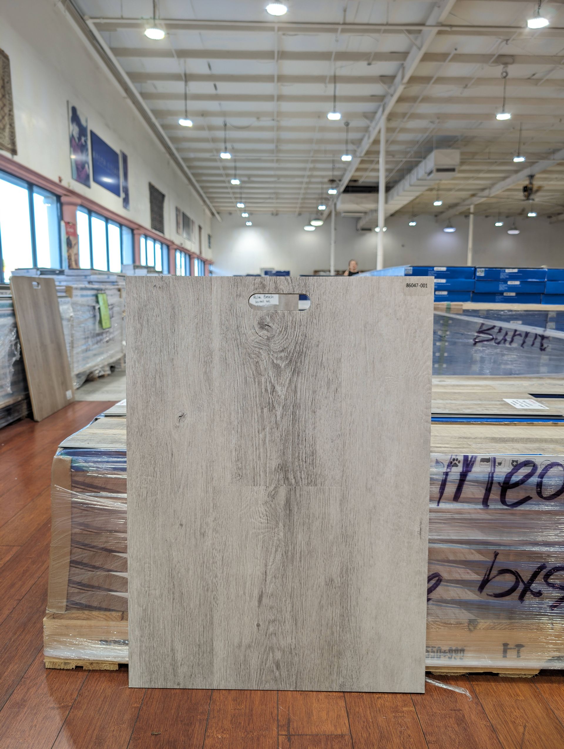 A light gray wood-grain sample tile in a warehouse setting.