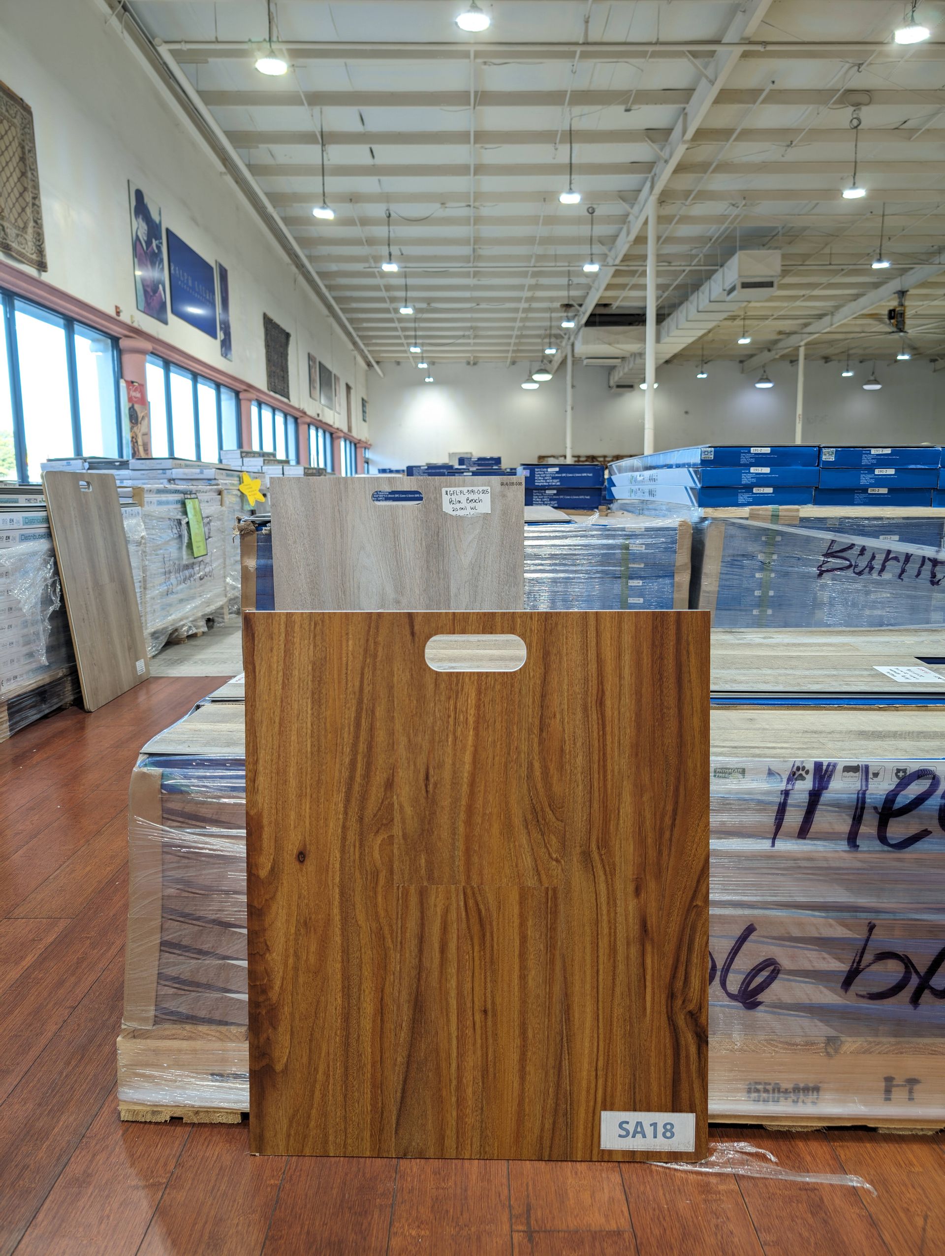 A wood-grained flooring sample with a handle stands in front of stacks of flooring in a warehouse.