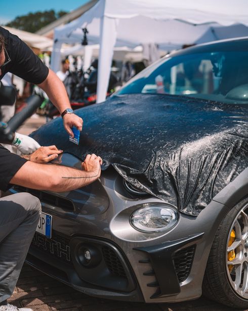 Persona che applica una pellicola protettiva al cofano di un'auto sportiva nera durante un evento all'aperto.
