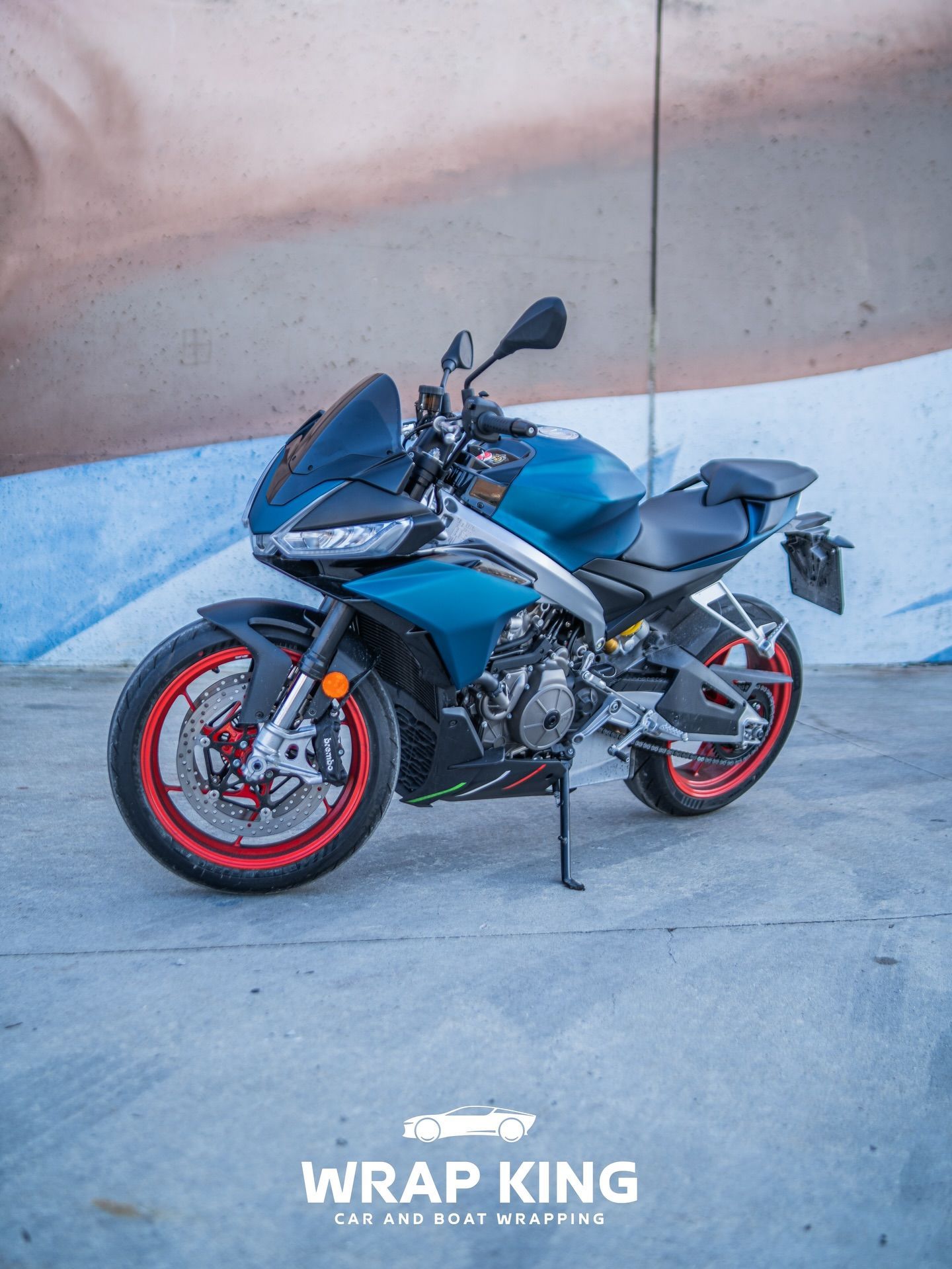 Motocicletta sportiva blu con cerchi rossi parcheggiata sul cemento accanto a un muro, vista laterale