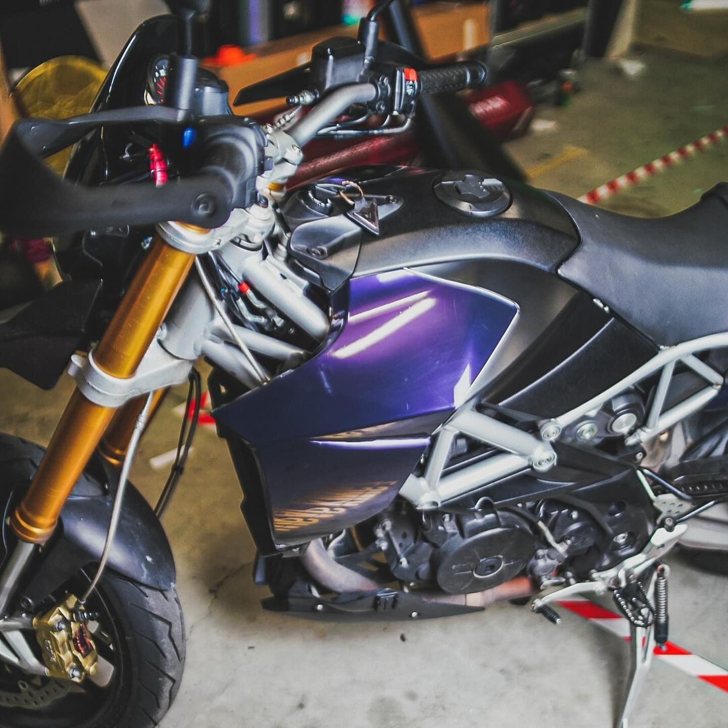 Motocicletta viola parcheggiata al chiuso, con manubrio, sella nera e telaio argentato in vista.