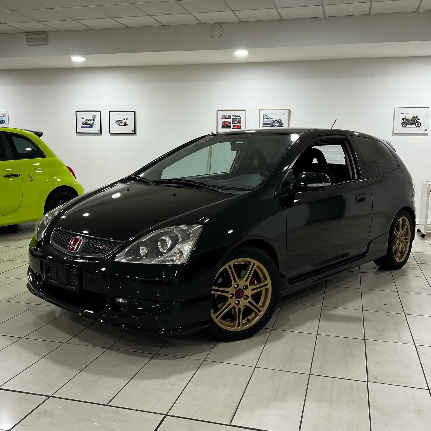 In uno showroom è esposta una Honda hatchback nera con cerchi dorati, sullo sfondo si vede un'auto verde lime.