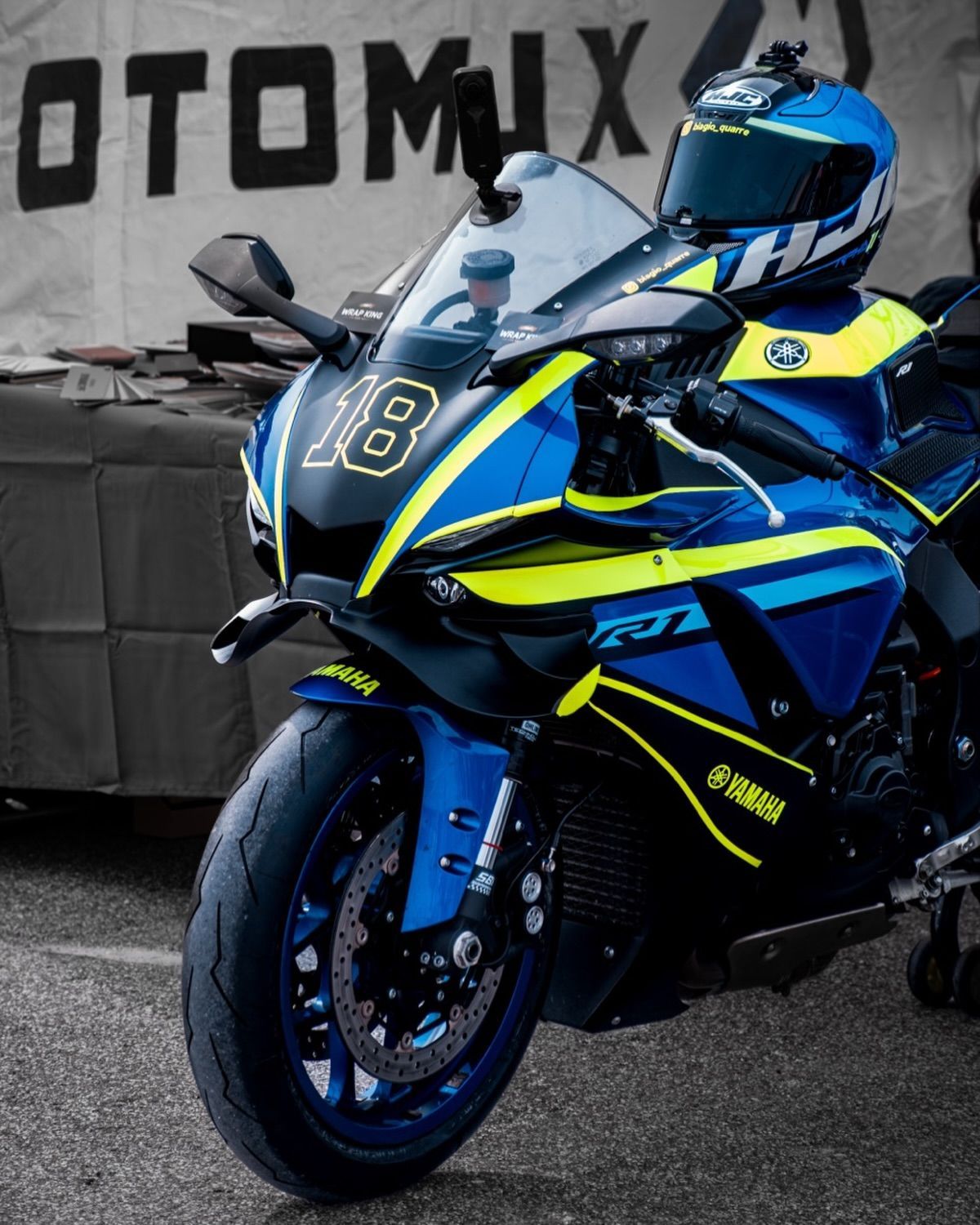 Motocicletta sportiva blu e gialla parcheggiata a un evento, con uno striscione nero sullo sfondo.