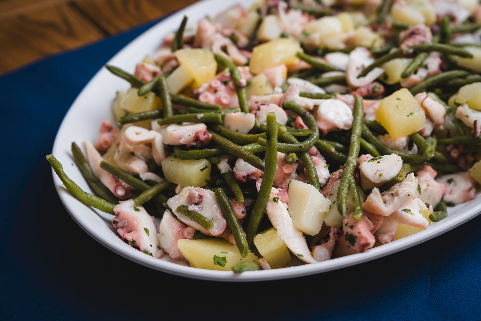 Piatto di insalata di mare con fagiolini, patate ed erbe aromatiche tritate su un tavolo blu scuro.