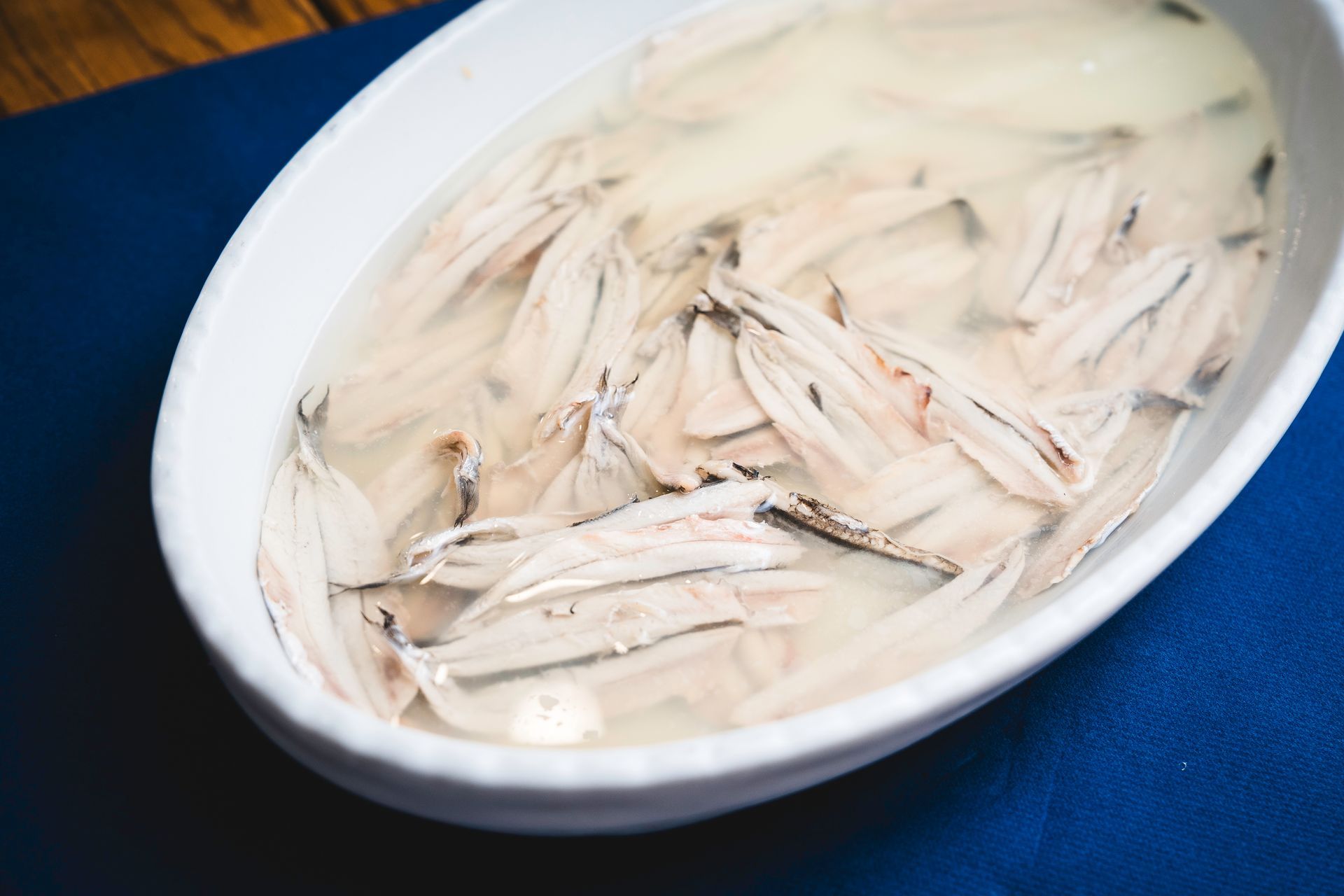 Una ciotola bianca contenente fette di pesce pallido immerse in un liquido lattiginoso su una superficie di stoffa blu.
