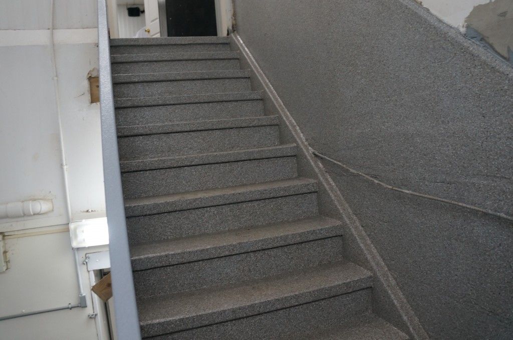 Escalier en pierre grise dans un bâtiment, avec une rampe à droite et une porte en haut.