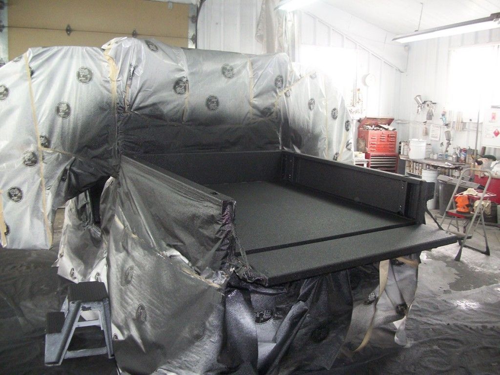 Dans un atelier, la benne d'un camion est recouverte d'un matériau protecteur noir. Elle est partiellement masquée par du plastique.