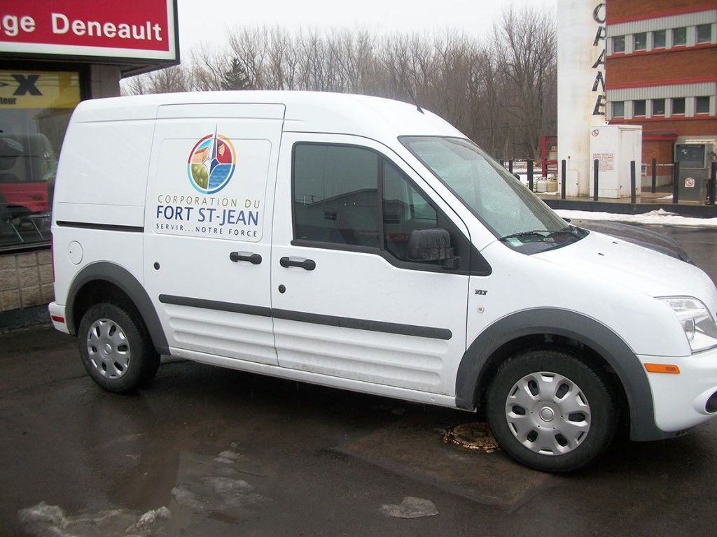 Une camionnette Ford blanche portant le logo de Fort Saint-Jean est garée devant un bâtiment.