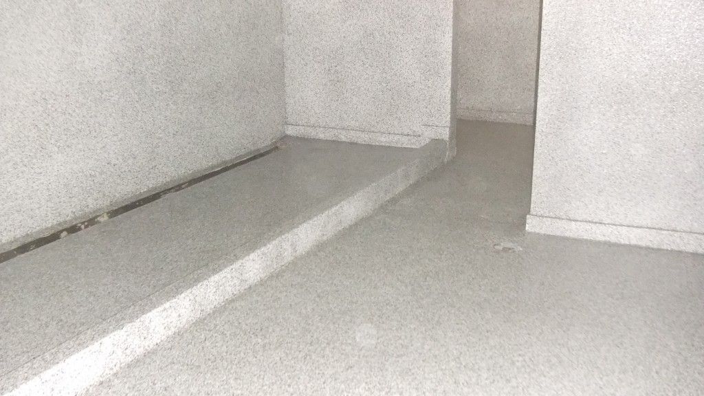 Pièce en béton gris, sol avec une marche donnant accès à un niveau supérieur. Les murs présentent une finition texturée.