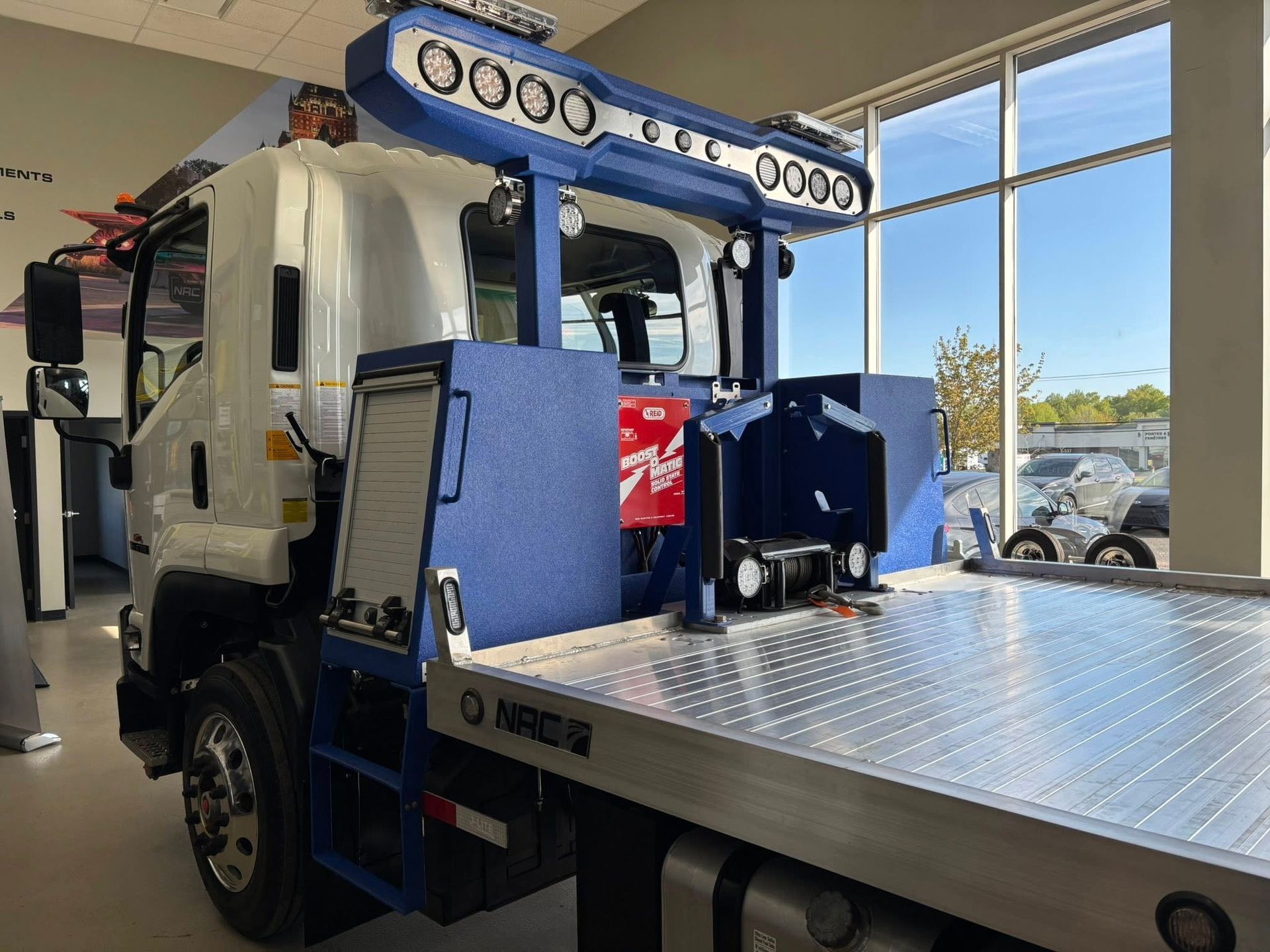 Camion de dépannage bleu et blanc à plateau allongé, stationné près d'une fenêtre à l'intérieur.