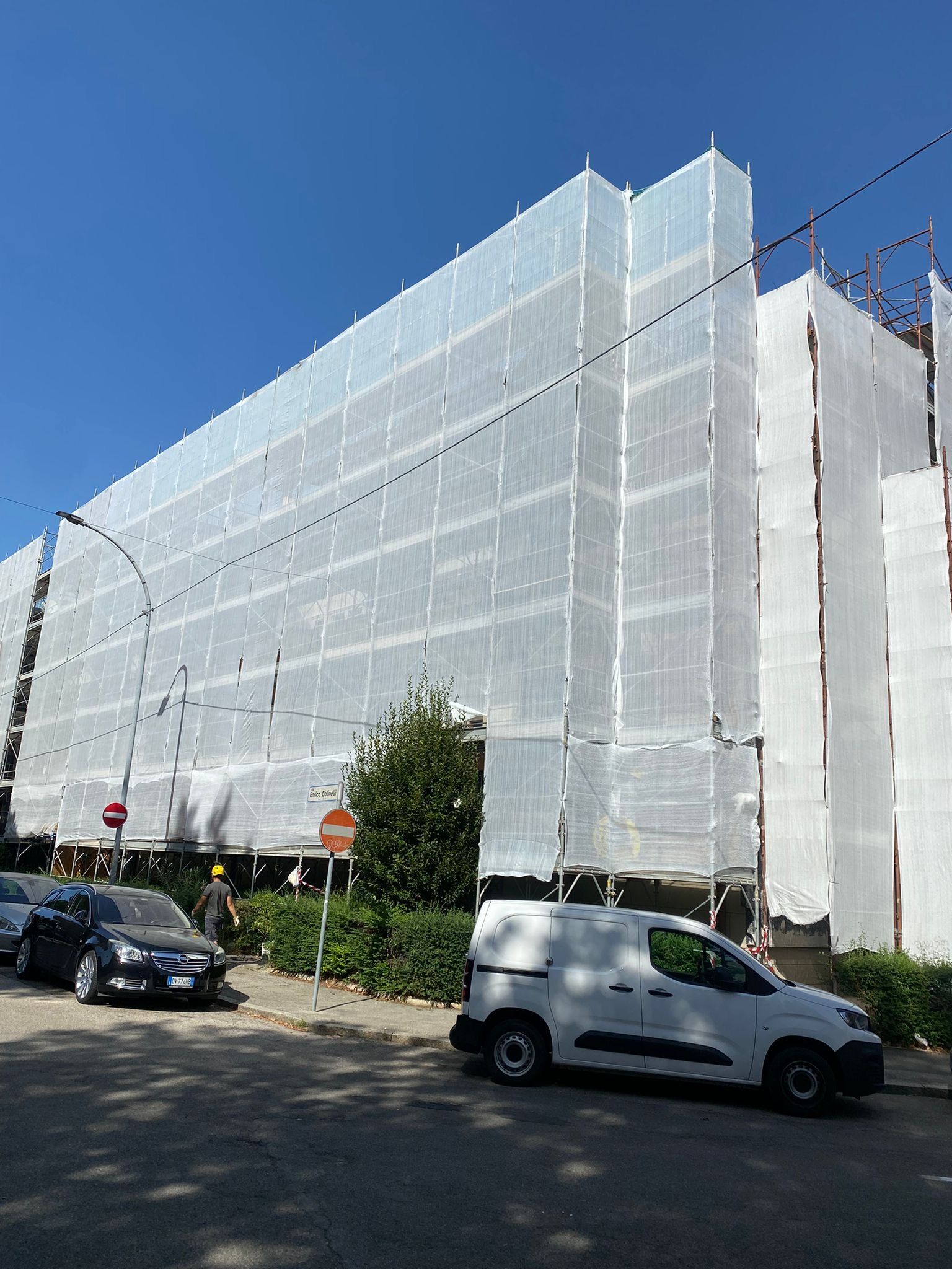 Scaffolding wrapped in white mesh around a large building on a city street with parked cars under a blue sky