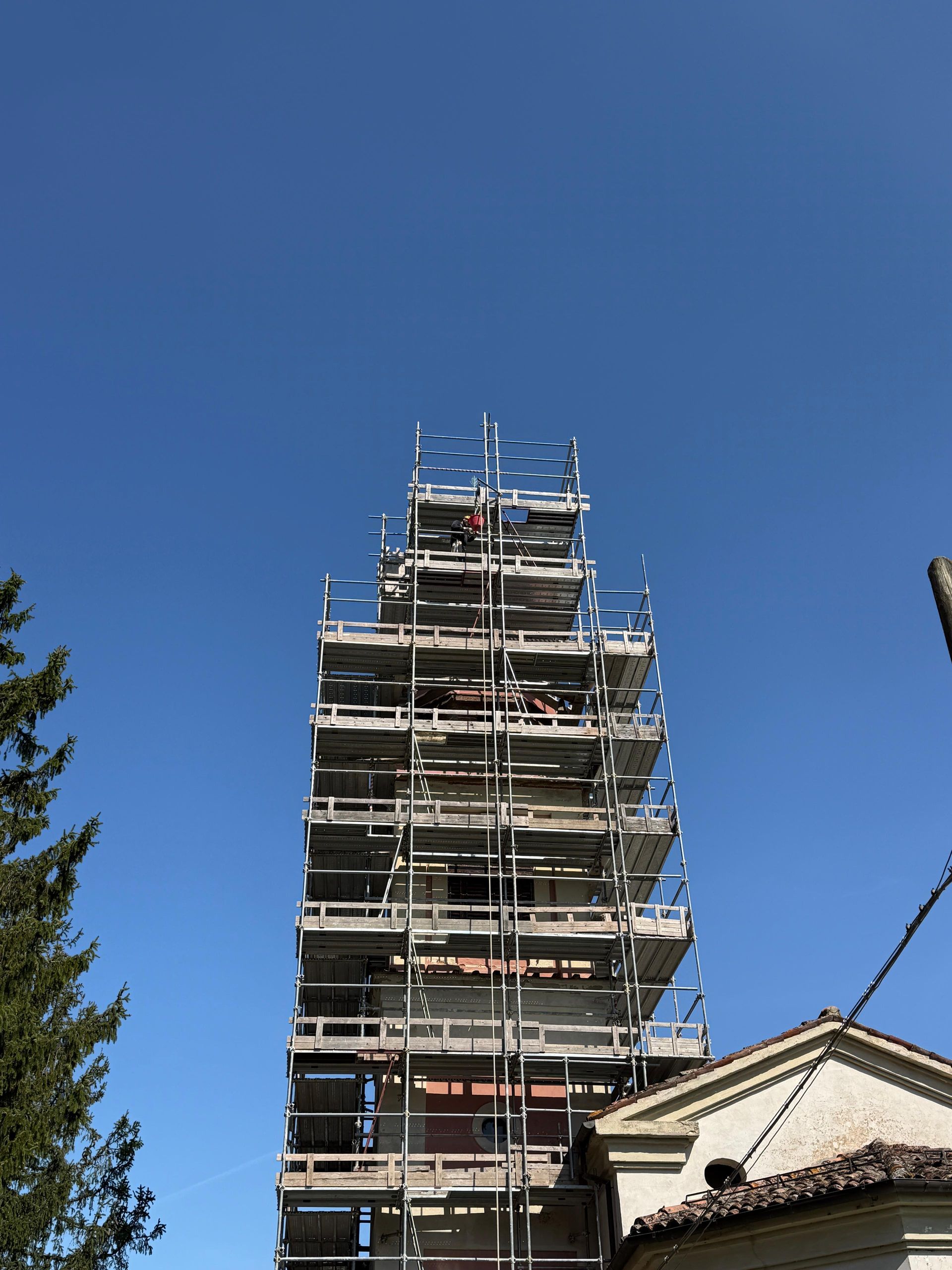 Scaffolded tower against a clear blue sky, with trees and a building roof in the foreground