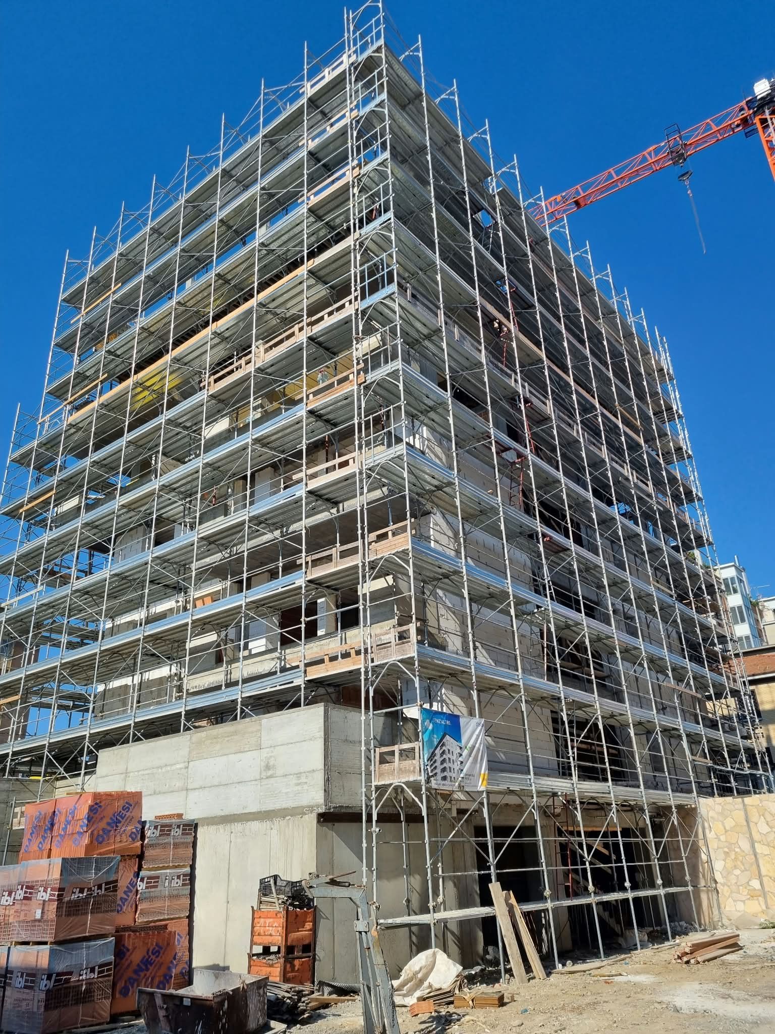 Scaffolding-covered building under construction against a bright blue sky, with a crane overhead