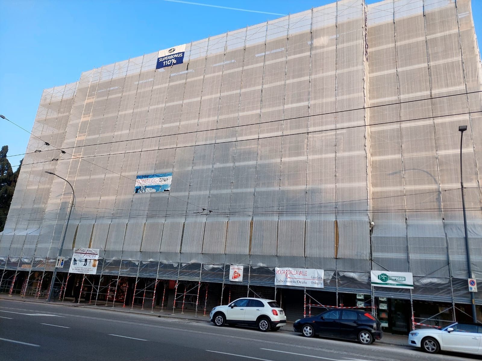 Large beige building covered in scaffolding and safety netting, with cars parked along the street below.