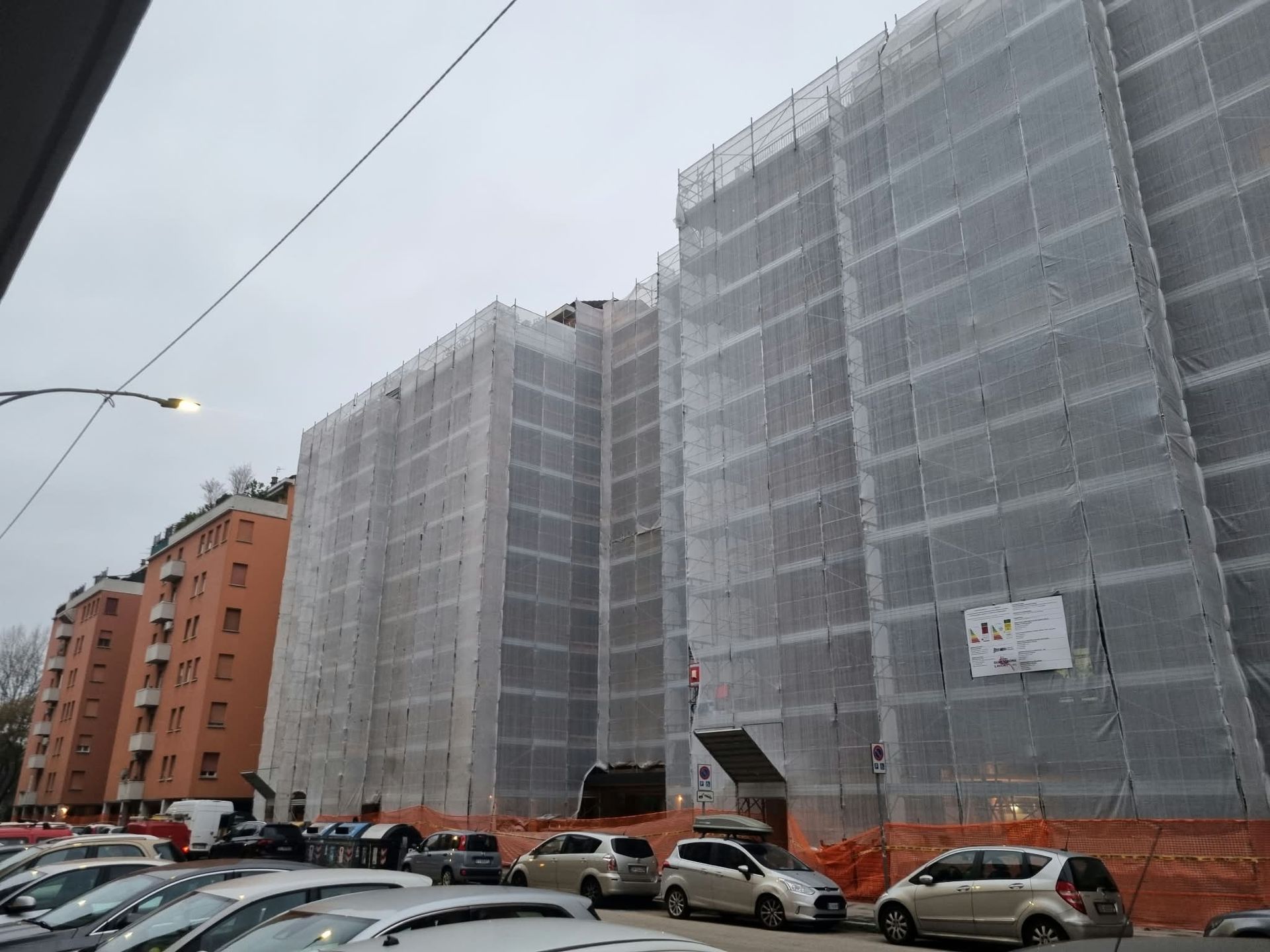 Apartment buildings wrapped in scaffolding and mesh, with parked cars along a city street on an overcast day