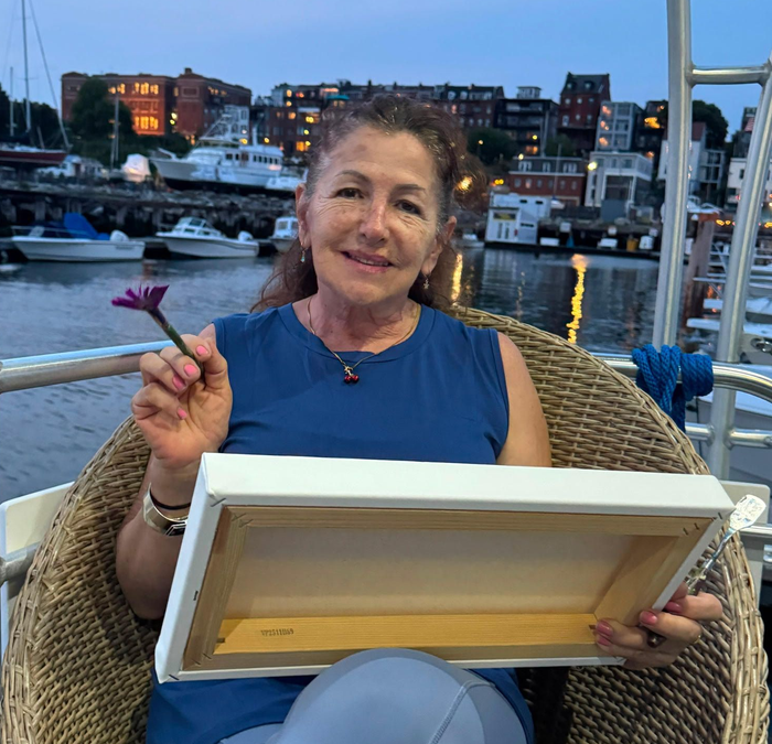 Woman smiling on a boat deck at dusk, holding a small purple flower and a framed canvas.