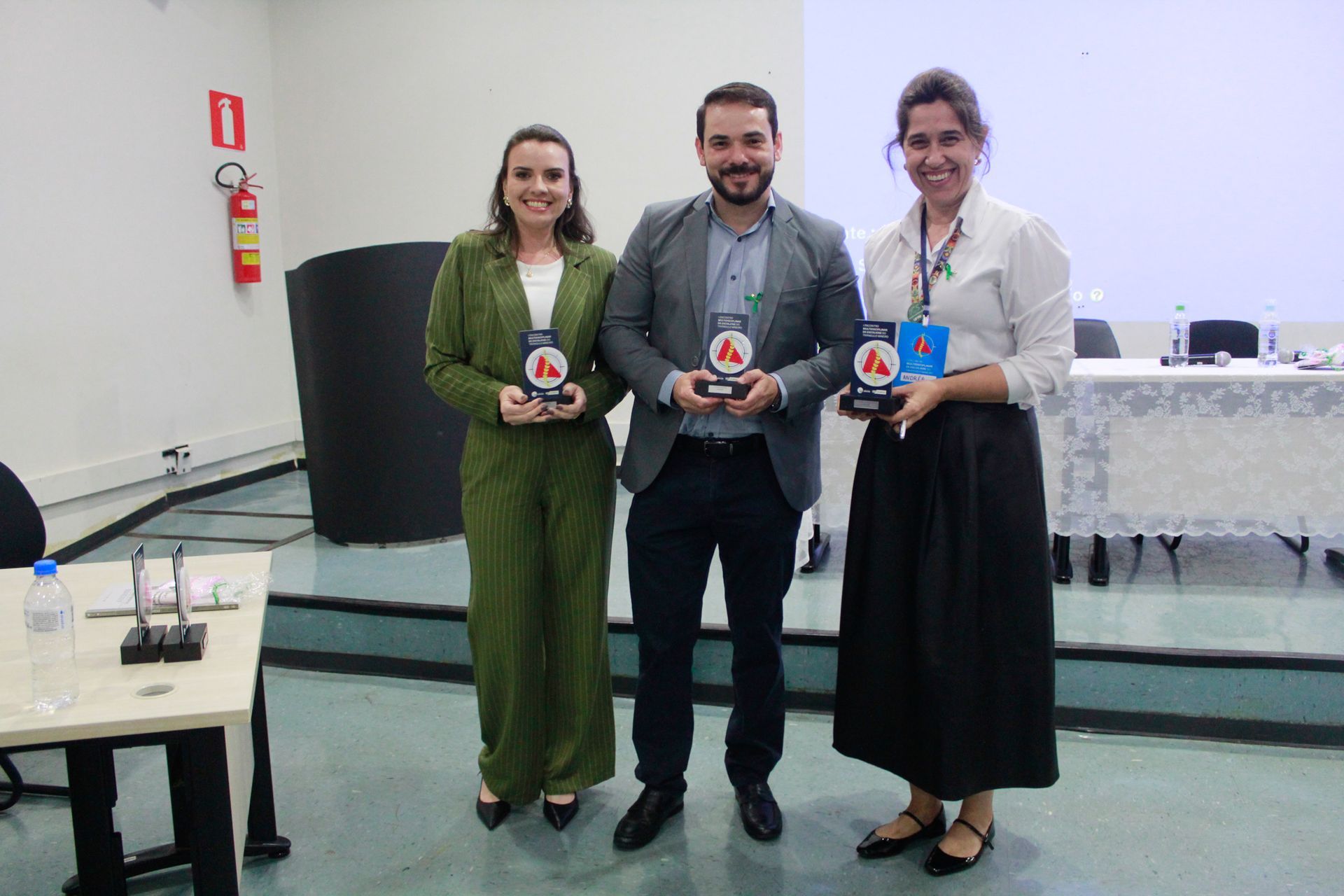Três pessoas estão de pé, uma ao lado da outra, em uma sala, segurando troféus.