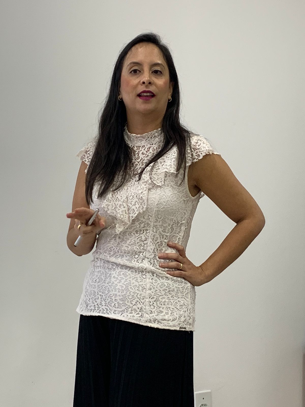Dra Francine com uma blusa de renda branca e saia preta está parada em frente a uma parede branca.