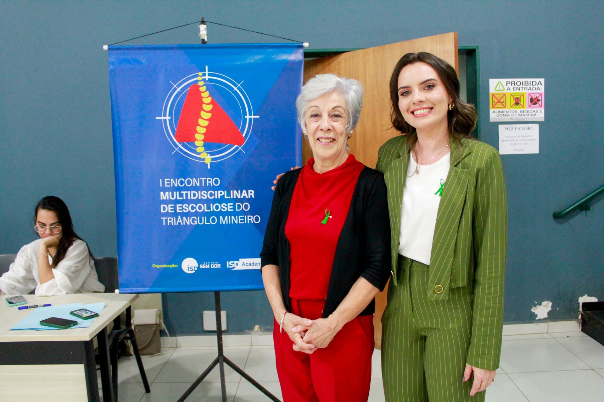 Duas mulheres estão de pé, uma ao lado da outra, em frente a uma faixa azul.