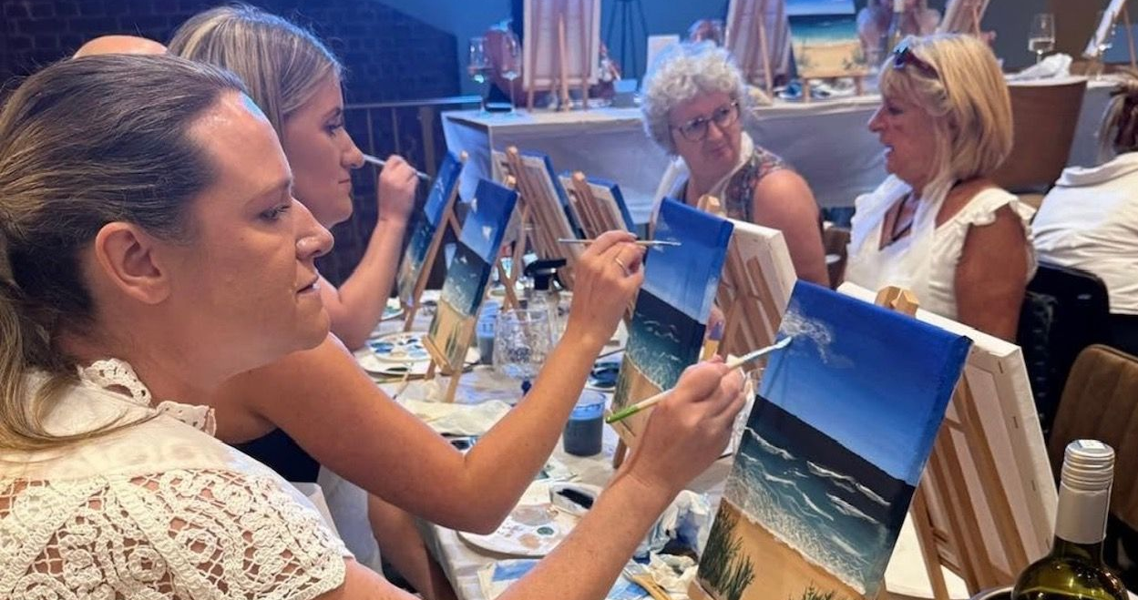 People painting at easels in a studio class, focused on seascape canvases