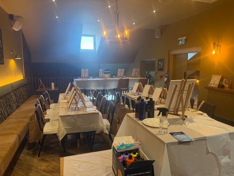 Banquet room with long tables set for an event, covered in white tablecloths and framed artwork on easels.