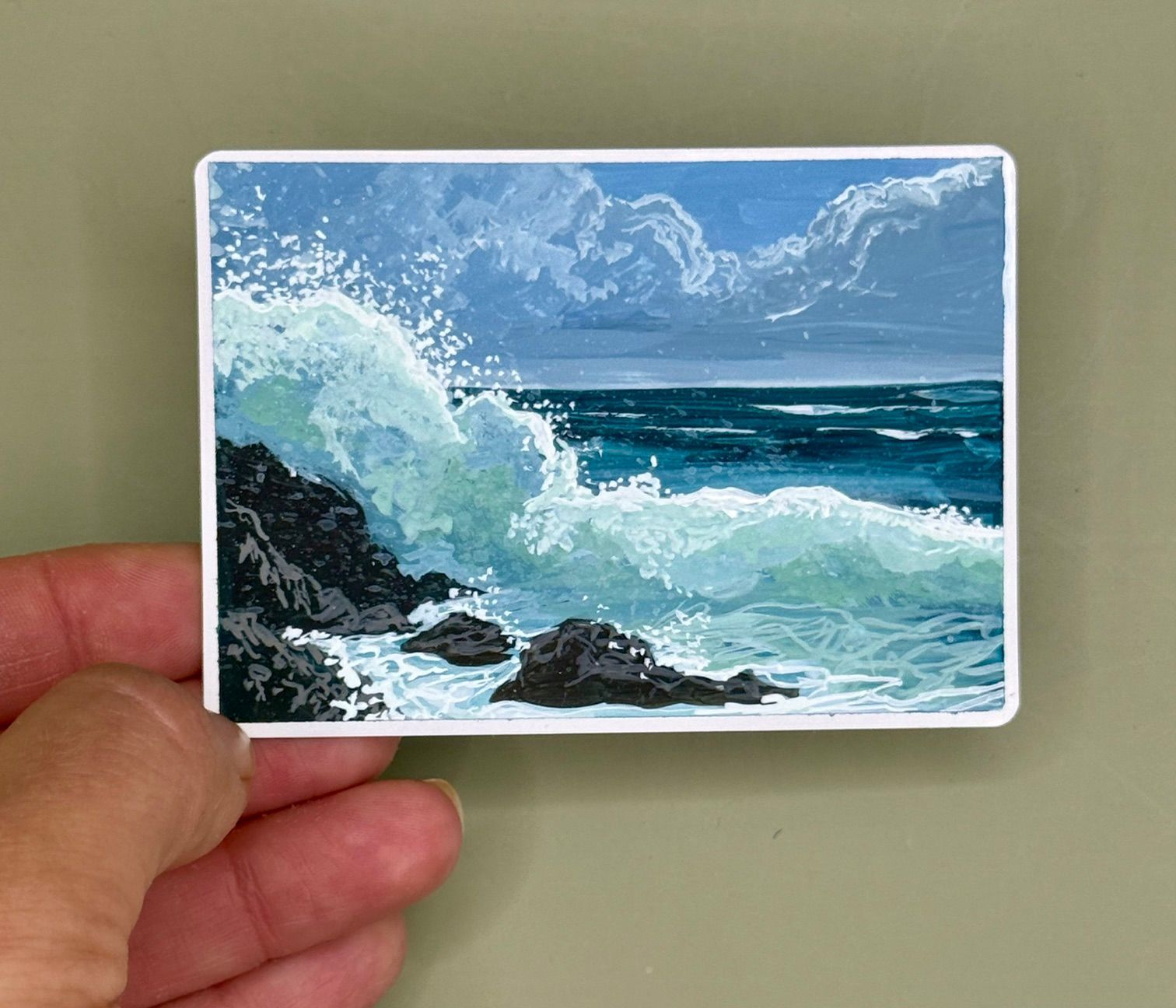 Hand holding a small card with a painted seascape of waves crashing on dark rocks