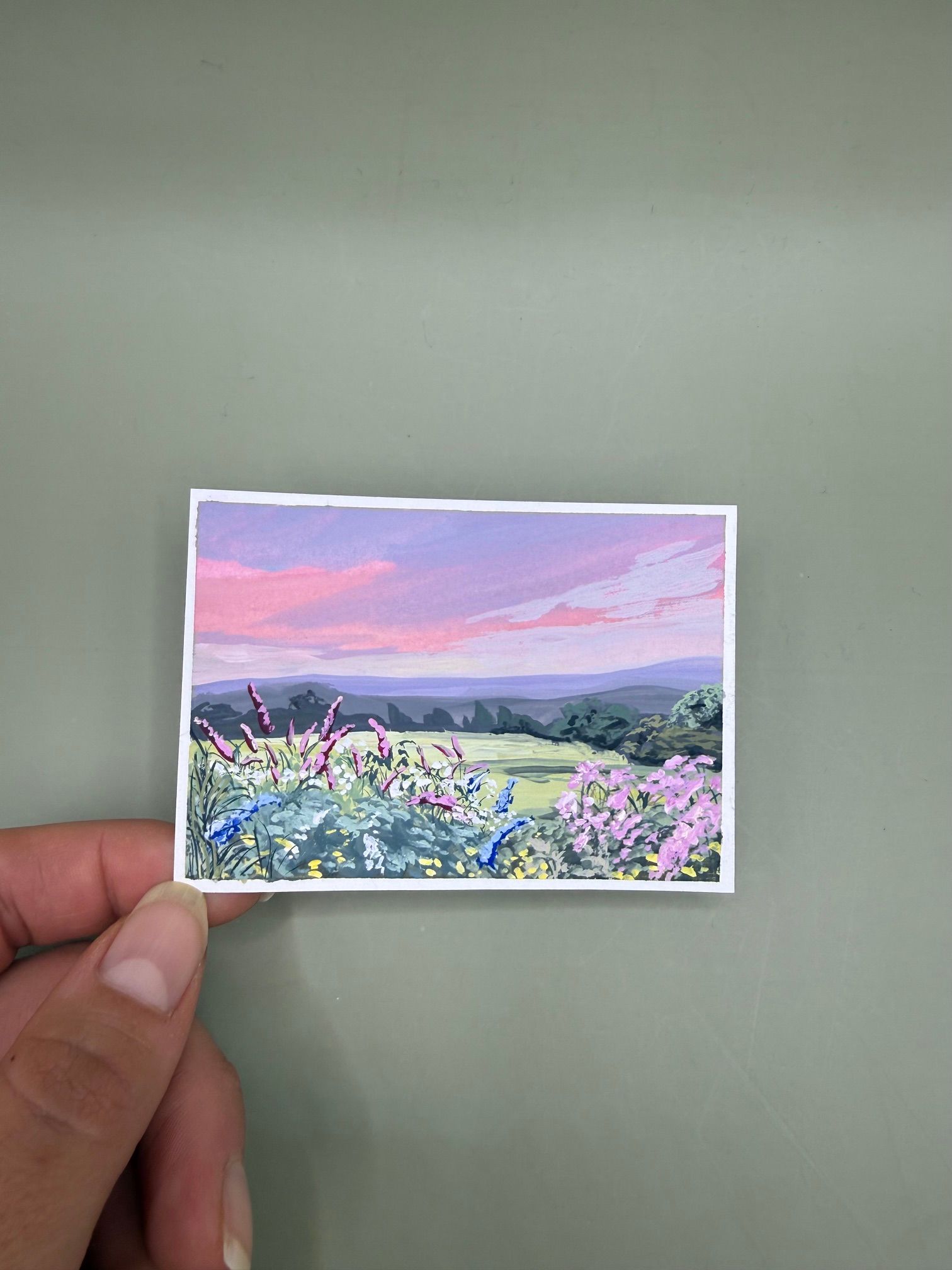 Hand holding a small landscape print of a flower field under a pink sunset sky