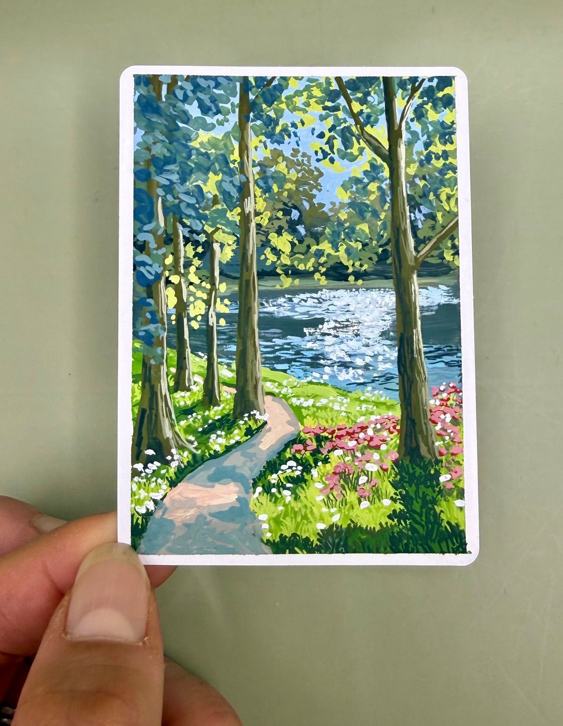Hand holding a postcard of a sunny lakeside path with trees and pink flowers