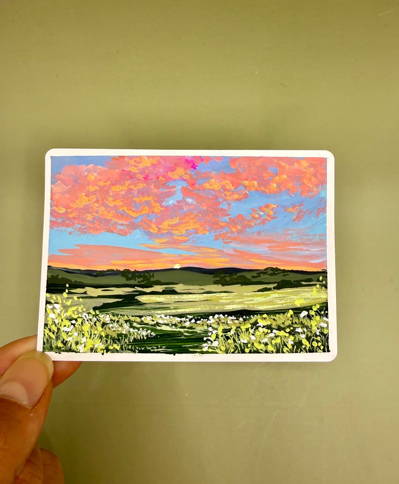 Hand holding a small landscape print with pink clouds over a green field and yellow flowers