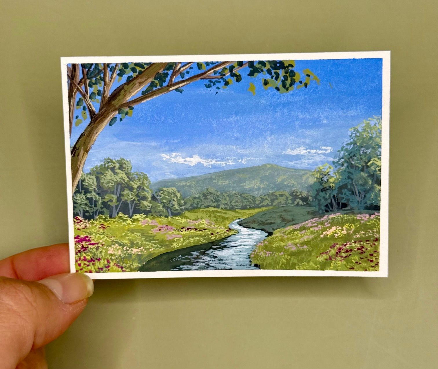 Hand holding a postcard-style landscape painting of a river, green hills, and blue sky.