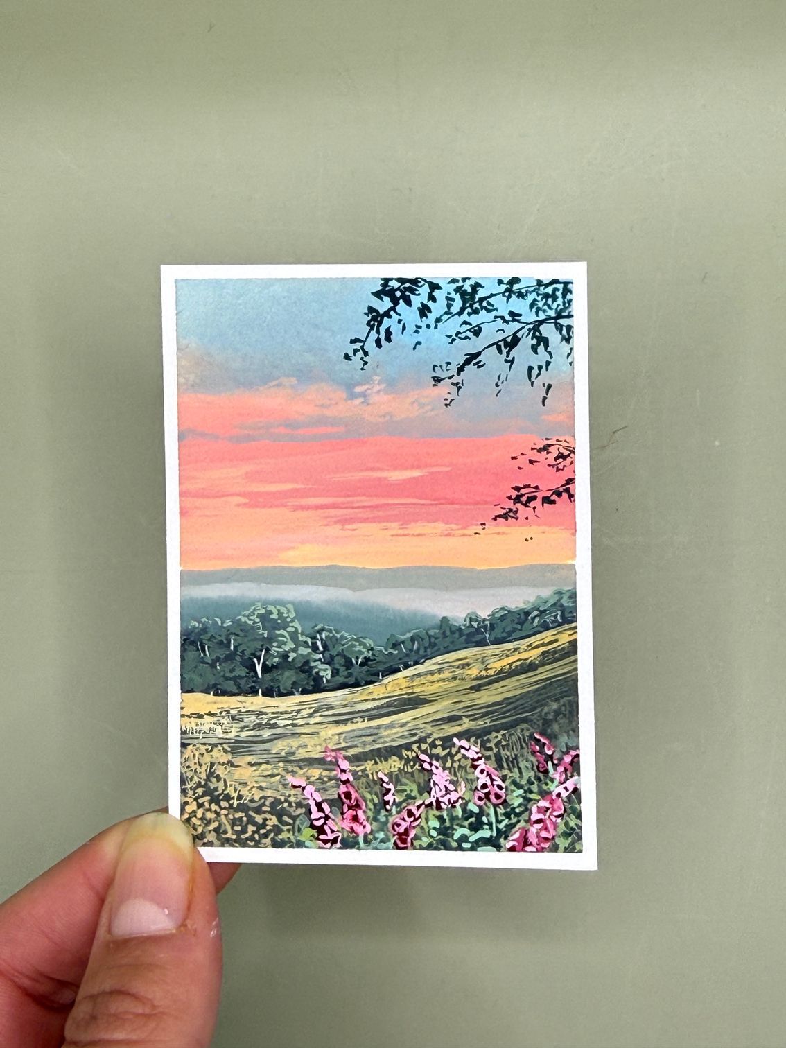 Handheld landscape print of a pink sunset over rolling hills with wildflowers and tree branches framing the view