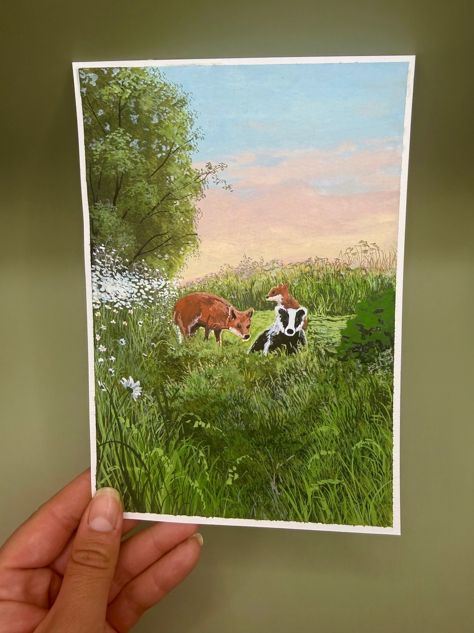 Handheld watercolor painting of a cow and a dog in a green field under a pastel sky