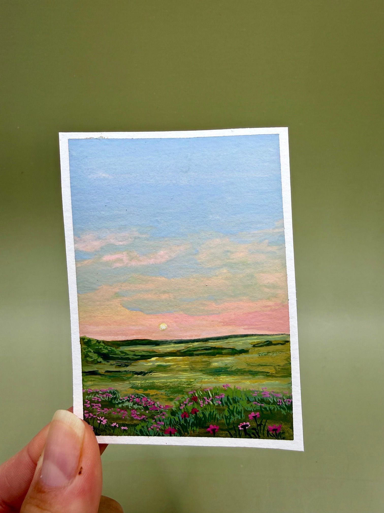 Hand-held miniature landscape print of a pink sunset over green fields and wildflowers