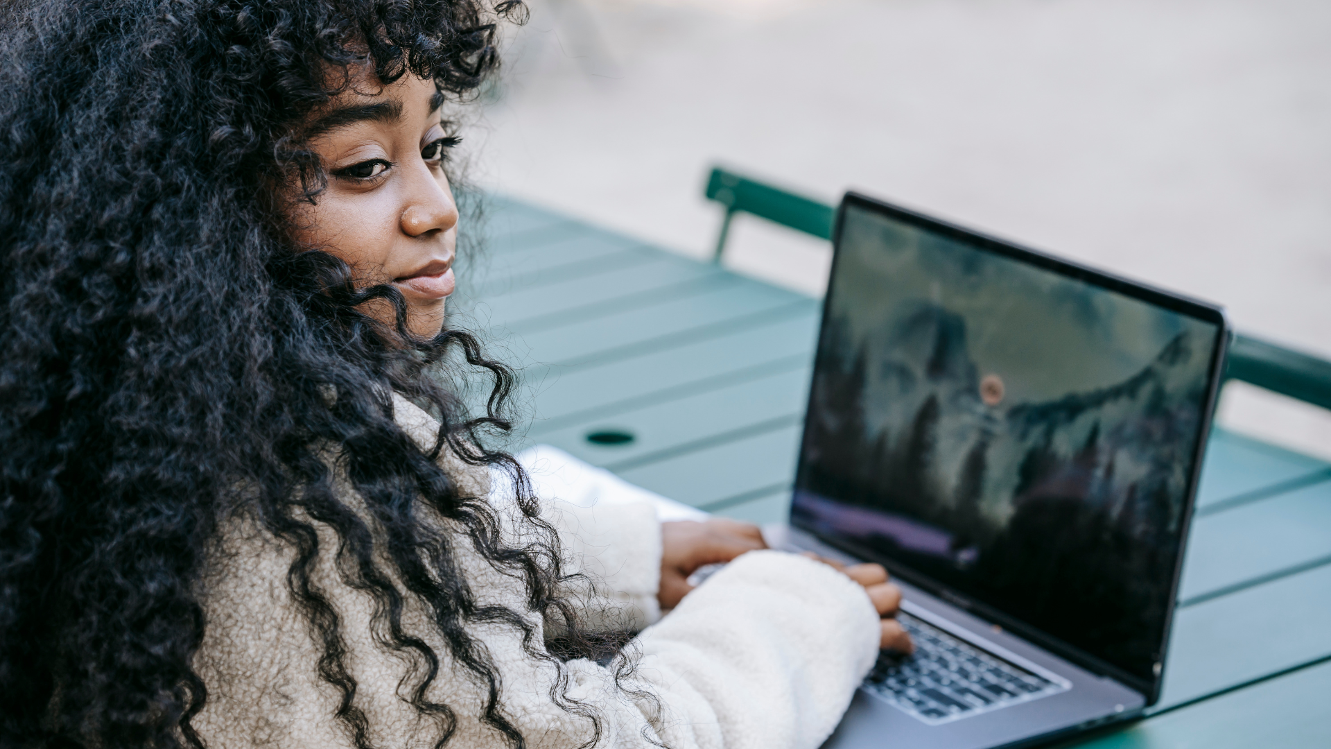 Person with curly hair using a laptop at an outdoor table, looking at the screen.