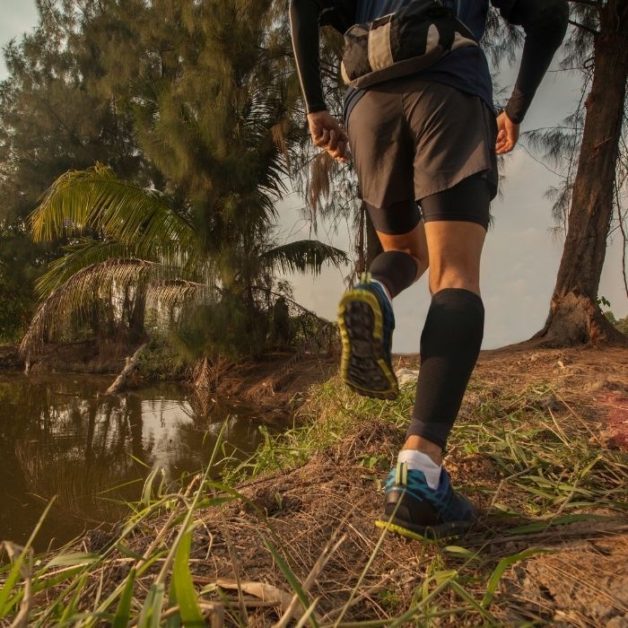 Runner in black compression socks and blue shoes jogging along a dirt trail beside a pond and trees.