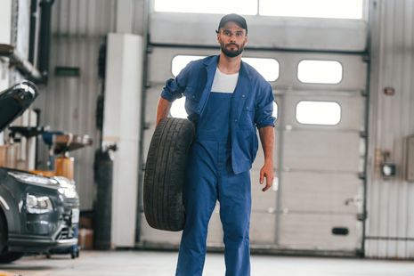 A man in blue overalls is holding a tire in a garage.