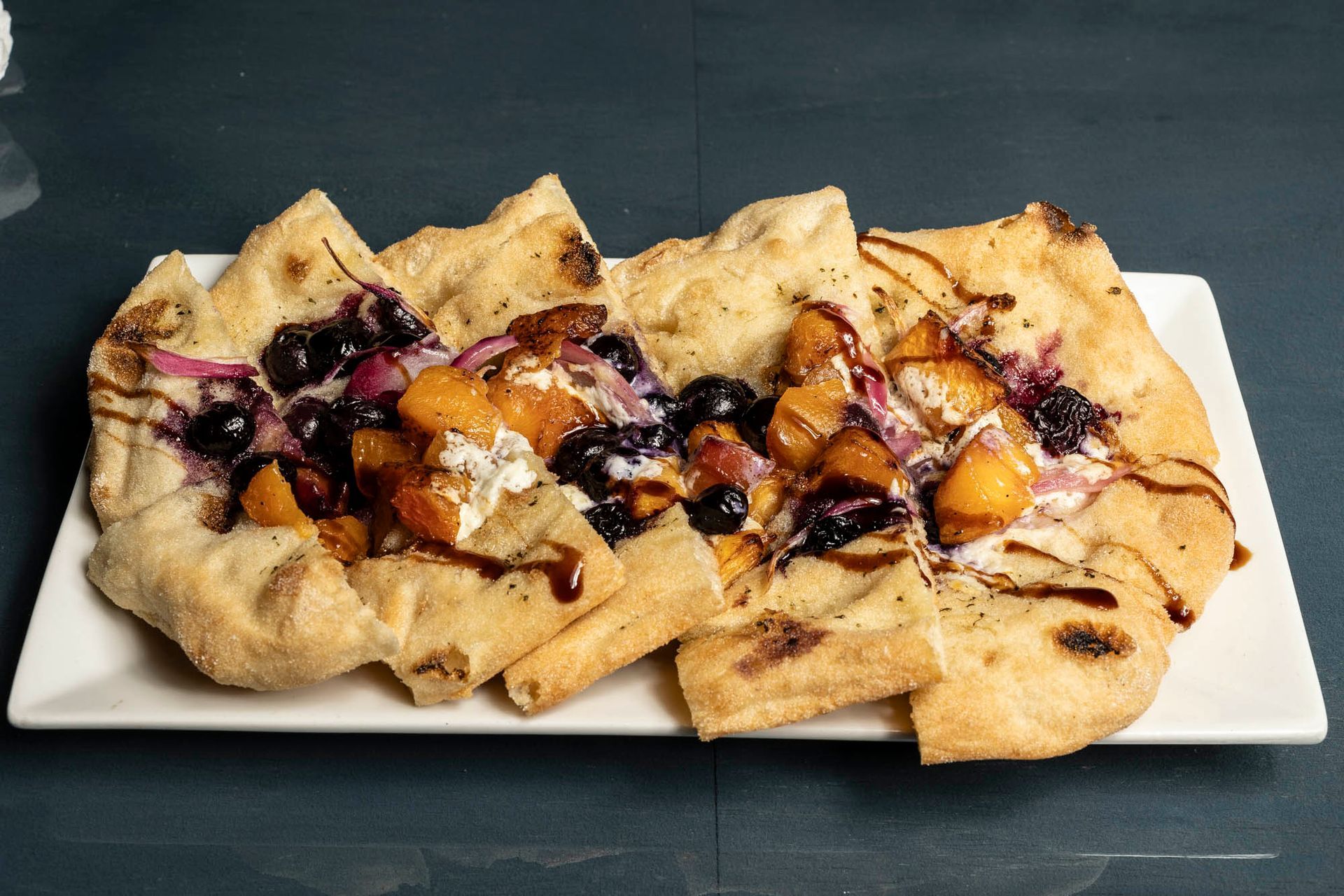 A white plate topped with a flatbread pizza with blueberries and peaches.
