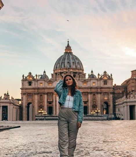 A person in a denim jacket and grey trousers standing in front of St. Peter's Basilica at sunset.